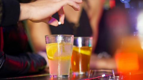 Two drinks on a bar. A hand is holding a tablet over one of the drinks. The photo is illustrative of a drink spiking incident.