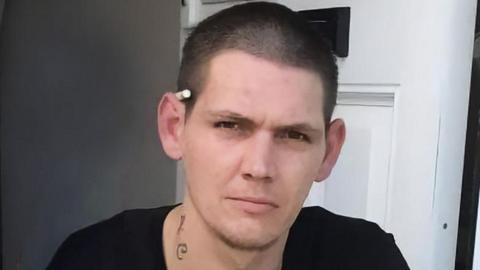A head and shoulders shot of a man with close cropped short black hair, a black t shirt and a cigarette behind his left ear, sitting on a doorstep