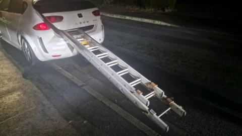 A white Seat Leon stopped on the side of the road, it has a large set of ladders sticking out the boot which is partially open.