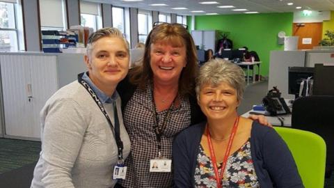 Three women looking at the camera and smiling. In the background is an office setting. There are desks with computers and storage. The wall in the background is green. There are windows on the left. 