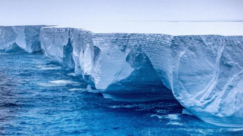 An edge of the A23a iceberg seen towering over the sea it is drifting through. The top of it appears perfectly flat and stretches into the horizon.