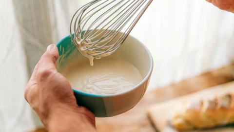 A person holds a blue bowl with a white sauce in it, and a whisk in the other hand