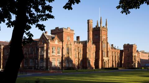 Queen's University Belfast, a large red brick building. Garden lawn out front. Tree in darkness in foreground.