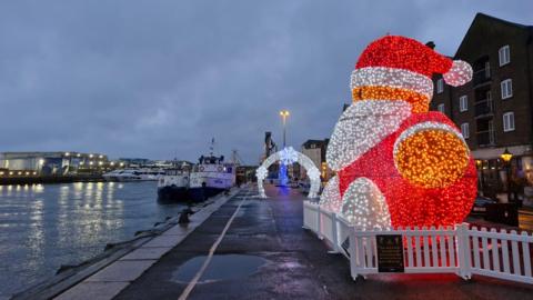 An LED Father Christmas alongside a coastal path with a harbour in the distance