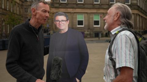 Aman holding a microphone and cardboard cut out of Andy Burnham interviews a man in a white shirt on a street