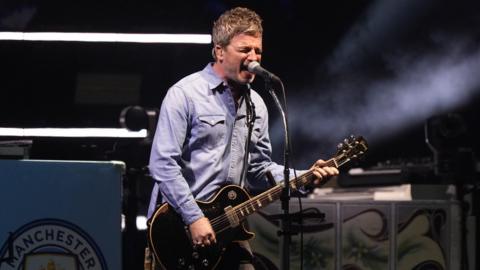 Noel Gallagher in denim shirt singing and playing a black Gibson Les Paul guitar on stage in Cardiff.