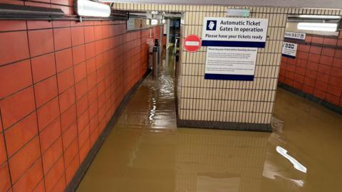 A corridor in a train station flooded with brown water. Ticket gates can be seen and the walls are tiled with red