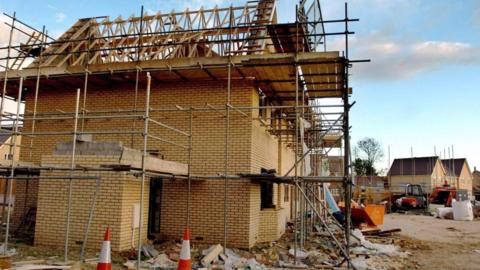 A partly built home, with scaffolding around it. Two red and white cones are in the foreground. Other part built homes and equipment is in the background. 