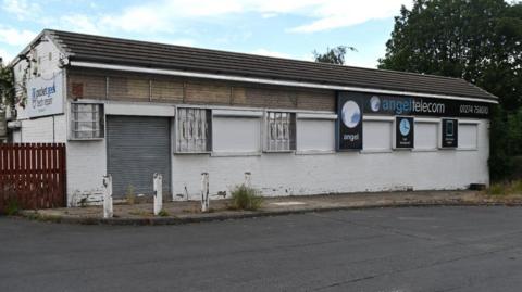 A rundown single-storey building with shutters over the windows and some panels on the walls missing.