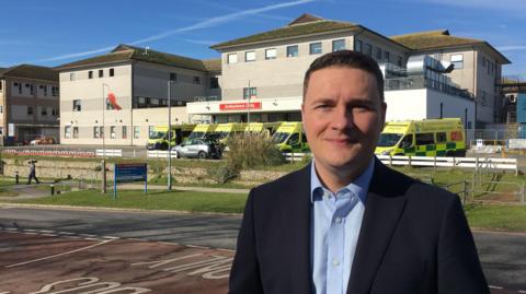 Wes Streeting is pictured outside the Royal Cornwall Hospital emergency department. He is wearing light blue suit shirt with a black blazer. Ambulances can be seen parked up in a row behind him.