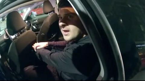 A man sat in the back sear of a black car is looking up at the camera, there is a shadow across his face and his mouth is open. He is wearing a black jacket.