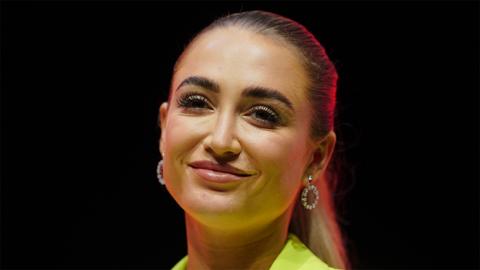 Georgia Harrison, a white woman, wearing round hoop earrings, smiling at the camera. She's wearing a green blazer, with the background all black.