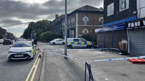 The junction of Yarm Lane and Lawrence Street in Stockton, with businesses including a vape shop and two-storey terraced homes. There are parked cars in front of some of the homes and along Yarm Lane which continues into the distance. A sign for Lawrence Street is in the right of the image. There is police tape, a police officer in uniform, a police car and a police van.