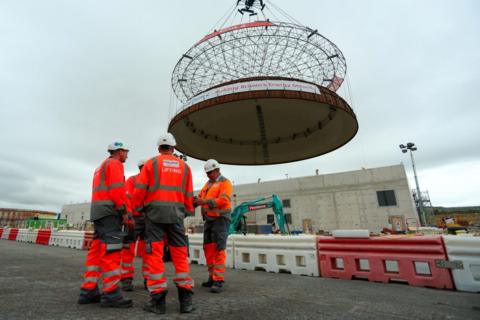 Work men in work jackets below a crane hoisting a ring of steel