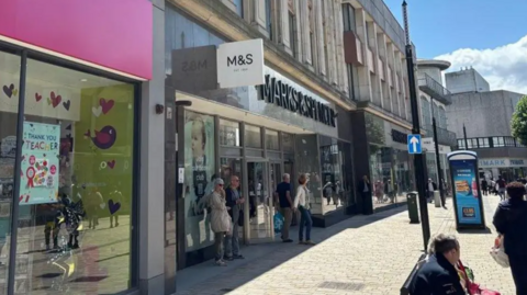 The store is on the high street in the city centre on the left of the image. There are shoppers coming out of the shop with other people standing or sitting in the pedestrianised street. The sun is shining.