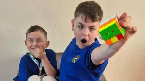 Two young boys wearing matching blue t-shirts. The younger boy is sitting in an armchair and giving a thumbs-up to the camera. The older boy is standing in front of the chair and holding up a Rubik's Cube in an heroic gesture. On a table in front of them are a selection of other toys, including several more Rubik's Cubes