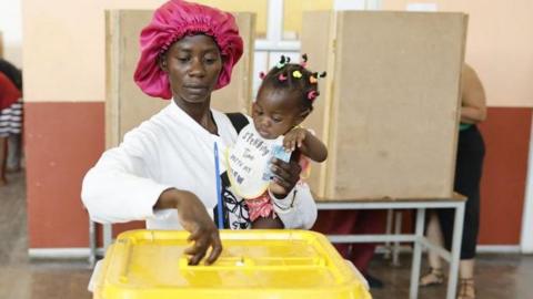 A woman wearing a pink head cap and cradling a baby puts a ballot paper into a ballot box with a yellow lid.