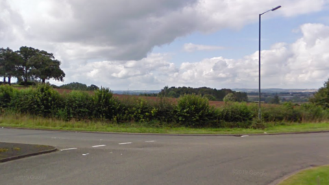 Street view image of a brown field with a green hedge along the perimeter at the side of a road junction 