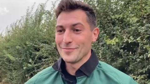 A man with dark hair is wearing a green top. Bushes are behind him.