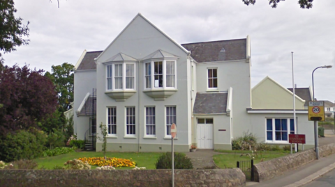 Google image of St Saviour's School in Jersey. The large grey building has big bay windows and a front garden with some bedding plants in it. There is a 20mph traffic sign on the pavement outside the school.