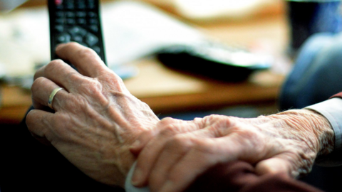A close-up photograph shows a person’s hands holding a television remote control while sitting indoors. Another remote and a mug can be seen out of focus in the background, creating a domestic home setting.
