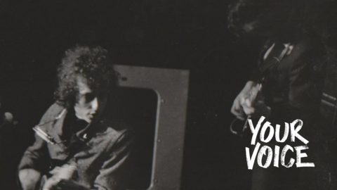 Bob Dylan with his distinctive curly hair, has his harmonica round his neck and is playing his electric guitar at Manchester Free Trade Hall