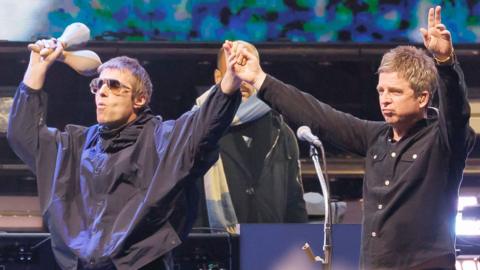 Noel and Liam Gallagher, both wearing black tops and jeans, raise their hands in the air on stage during their reunion tour on stage. Liam is holding maracas in his right hand.