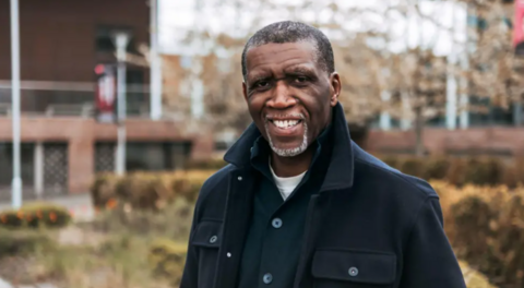 A man with short dark hair, a beard and a large dark blue coat standing outdoors with the blurred shape of a building behind him