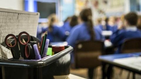 A box of stationery - including scissors and pencils - with primary school pupils in the background.
