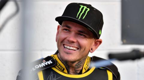 Tai Woffinden sits at the side of a speedway circuit and smiles as he talks to someone out of view. He is wearing black and yellow race overalls and a black baseball cap with a green logo on the front.