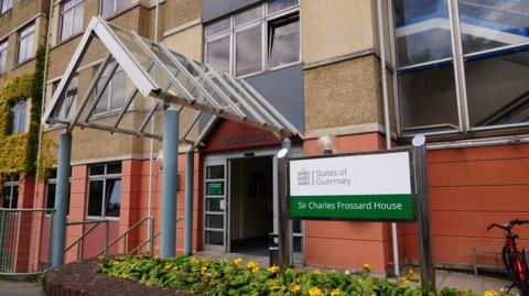 A green and white sign which says Sir Charles Frossard House, underneath the States of Guernsey logo. To the left of the sign is an automatic doorway with an arched covering above a reception sign. 