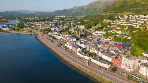An aerial view of Fort William on a sunny day. There are rows of houses and other buildings along the shore of Loch Linnhe.