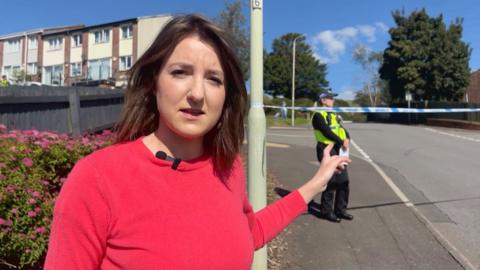 Reporter Lucy Vladev, a young woman with brown hear and a pink jumper stands in front of a police cordon on a residential street, with a police officer standing behind the cordon