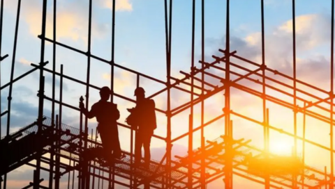 Two builders stand on scaffolding as the sun sets behind them on a construction site.