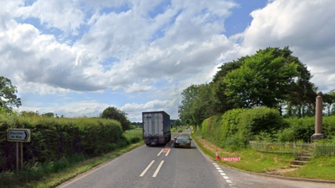 A main road with traffic travelling both directions, with a village road sign to the left, and a stone war memorial to the right