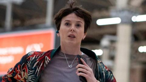 Alexandra Roach as Abby running through a train station wearing a grey T-shirt and patterned jacket