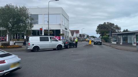 A police officer talks to a driver in a black car as colleagues further up the road deal with a car crash. Other vehicles including a silver Porsche and silver van are on the road. An M&S Food shop in in the background.