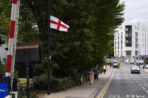A St George's flag flutters in the wind on the Isle of Dogs, east London. A woman wearing a head covering is walking on the pavement with a shopping bag and someone in front of her is pushing a pushchair. 