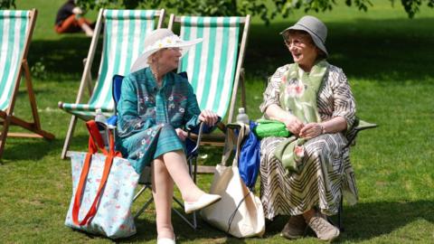 Two women wearing hats and print dresses sit in a sunny park.