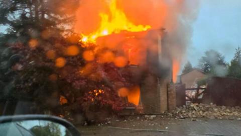 A blurry photo through a wet car window of a burning house, with all the windows and doors blown out and piles of brick rubble next to it. The building has orange flames shooting out.