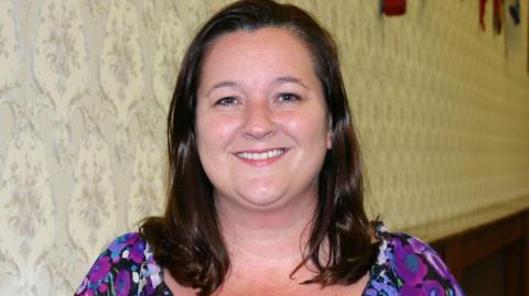 Marion Atkinson has long brown hair and is smiling. She is wearing a patterned purple and black dress.