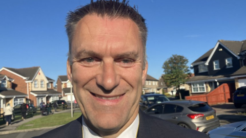 A smiling man with dark jacket and white shirt. He is standing in front of some modern houses with cars parked outside. There is a blue sky and the sun is shining