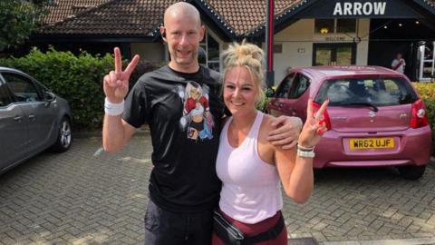 Giles and Katy stood side by side making peace signs with their hands and smiling at the camera. They are stood outside in a pub car park in running gear.