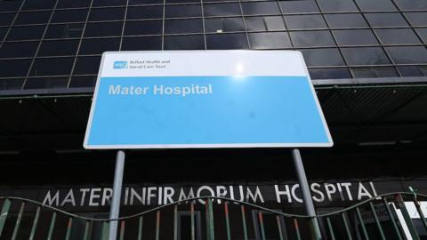 Front of the Mater Hospital. Green wavy railings are in the foreground and a sign reading 'Mater Infirmorium Hospital' in silver letters is behind. Another blue sign with white text says 'Mater Hospital'. Dark windows are above. Photo taken from below.