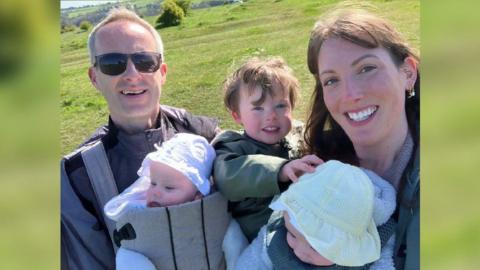 Lorna and Paul Jenkins smile with their three children - Joe, Lily and Ruby - in a selfie on a sunny day. The family of five are on a walk through a field. Lorna is holding Joe while she has one of the girls in a front carrier, and Paul has the other twin in a front carrier, too. Lorna has long brown hair with curtain bangs, while Paul has short grey hair and sunglasses on. Joe is wearing a green coat, and Lily and Ruby are wearing floppy cotton sun hats.