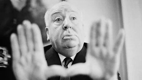 Alfred Hitchcock. It is a black and white photo and he his holding his hands up to the camera