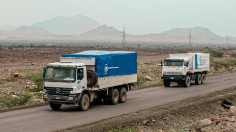 Two trucks bearing the World Food Programme logo travel on a road in a remote-looking area.