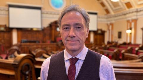 Carl Edwards, a man wearing a light-coloured shirt and a dark-coloured waistcoat, is standing in a council chamber. He is smiling for the camera.