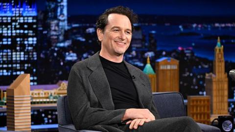 Matthew Rhys, a man with dark-brown hair, wearing a black shirt and greyish blazer is smiling with his hands rested on his leg. The background shows an image of black and orange buildings in the dark. He appears to be a guest on a chat show.