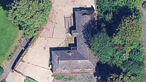 Image from above of an L-shaped building with hardstanding and trees around the building